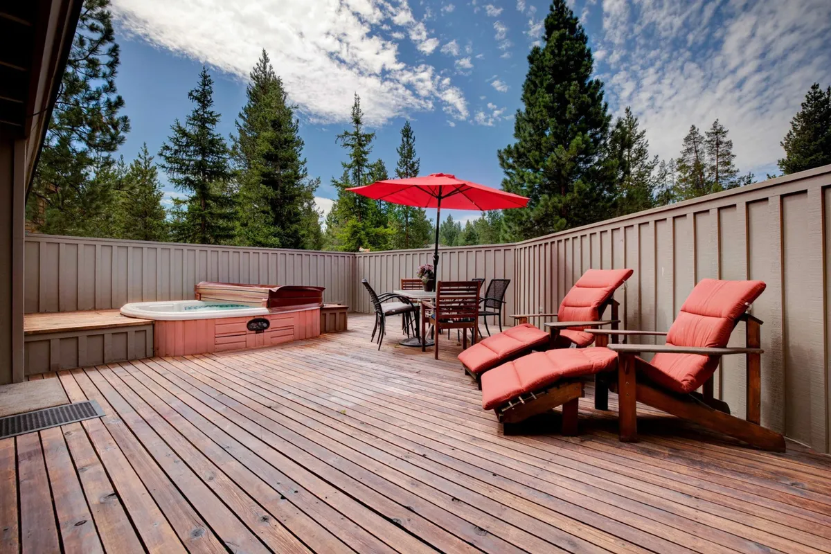The image showcases the home's inviting wooden deck at sunset, featuring a private hot tub under a red umbrella, comfortable lounge chairs for relaxing or sunbathing, and a spacious outdoor dining table perfect for shared meals. The tranquil atmosphere suggests evenings spent unwinding in the hot tub or gathering with friends and family, all surrounded by the serene Sunriver setting.