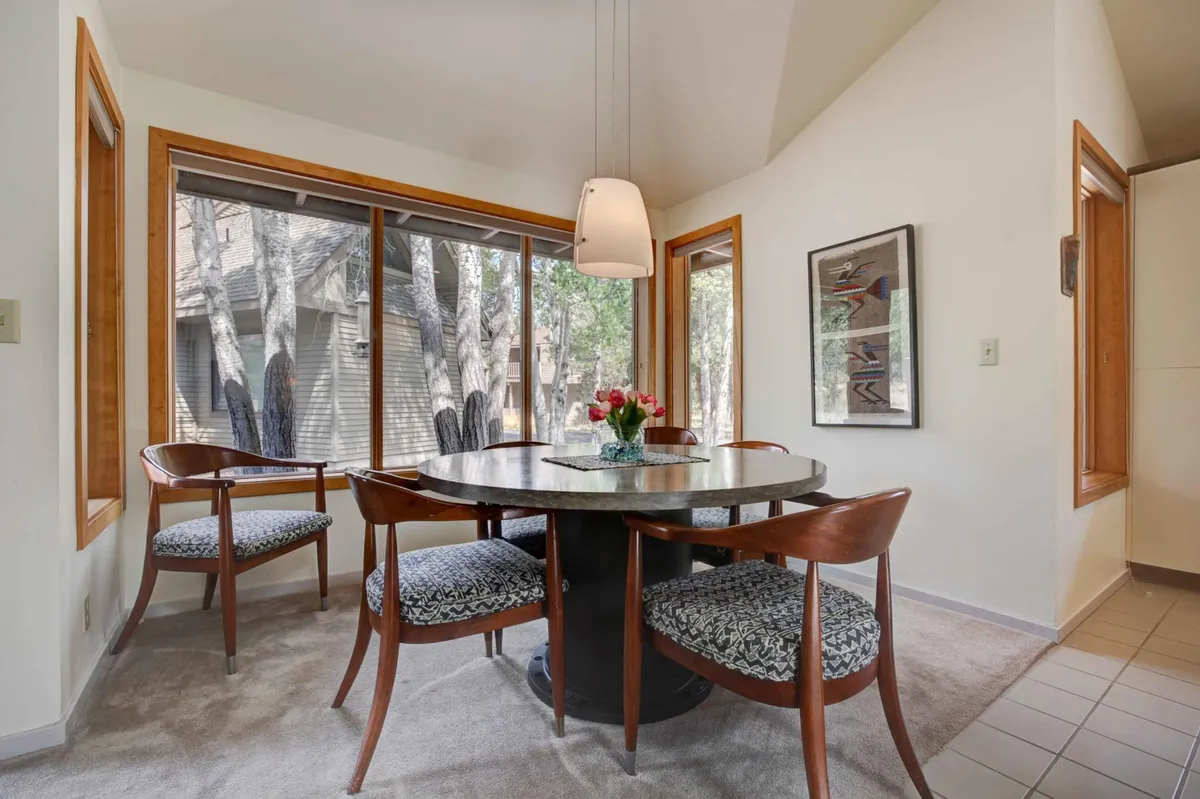 The dining area features a round table with stylish chairs, a flower vase centerpiece, and expansive windows offering beautiful natural views. The space is filled with sunlight, evoking warmth and inviting guests to enjoy meals amid a bright, cheerful ambiance.