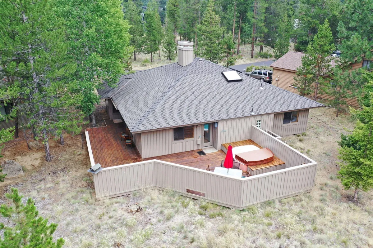 A wooden deck featuring a bubbling hot tub shaded by a red umbrella, surrounded by lush greenery. The scene is tranquil, with seating nearby for outdoor dining and plenty of space to unwind while immersed in nature. The view suggests a peaceful retreat, ideal for relaxing after a day of activities.