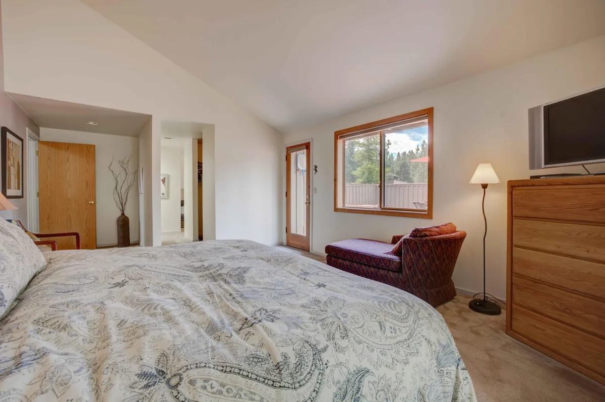 A serene bedroom with a bed featuring patterned bedding, an armchair nestled by a large window with outdoor views, a wooden dresser, and a floor lamp. The space is filled with calming natural light, ideal for relaxing after time outside.