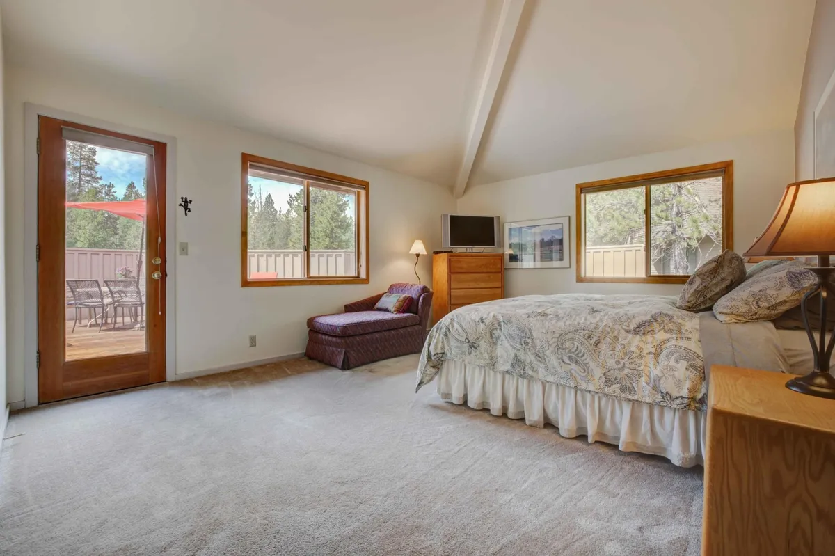 A spacious, sunlit bedroom featuring a large king bed with decorative bedding, a comfortable chaise lounge, a wooden dresser topped with a flatscreen TV, and expansive windows letting in plenty of natural light. The room exudes serenity and offers a private, restful atmosphere—ideal for unwinding anytime of day.