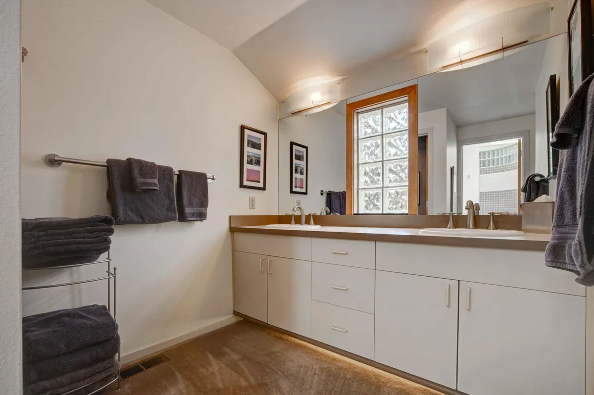 A bright, modern en suite bathroom featuring a double sink vanity, large window for natural light, framed photographs, and a towel rack. The space feels tranquil and ideal for self-care routines or unwinding after a long day.