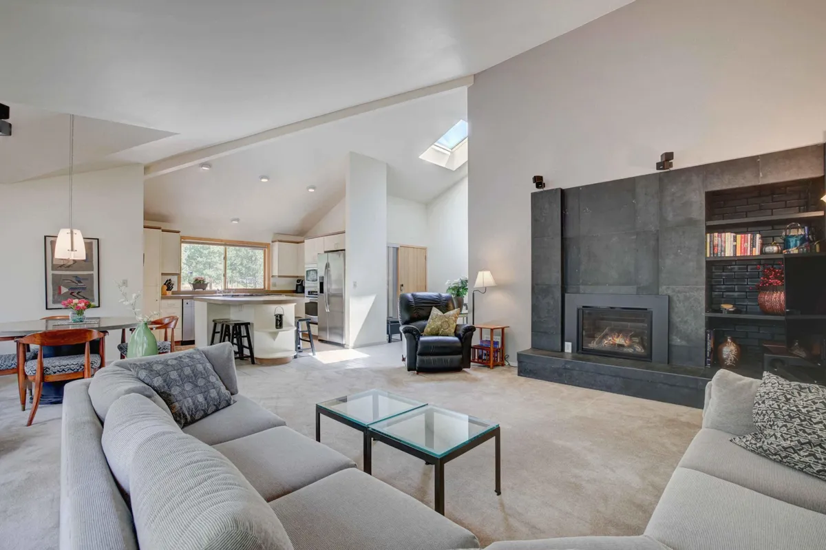 This image captures a spacious open-concept living area featuring a plush sectional sofa arranged around a dramatic dark marble gas fireplace. Generous sunlight streams in from large windows, while the kitchen and dining spaces are visible in the background, emphasizing the room's social and connected layout. The atmosphere is inviting, with comfortable seating and stylish, retro-inspired décor highlighting the space's 80s mod aesthetic.