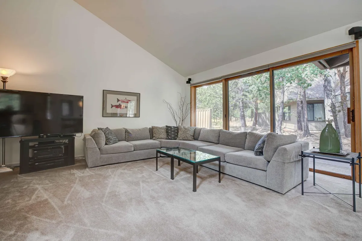 A comfortable common area with a large sectional sofa faces a glass coffee table and an oversized flatscreen TV. Natural light pours in through wide windows, revealing a serene view of the outdoors. The space is designed for relaxation and group gatherings, echoing the living room's function as a vibrant social hub.